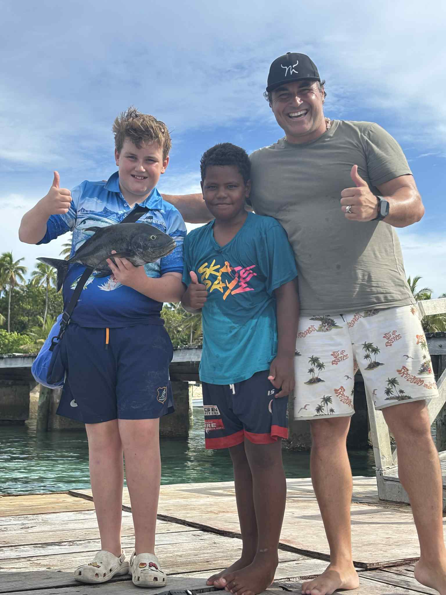 Family on a wooden jetty with a freshly caught trevally, palm trees behind